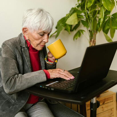 An elderly woman in a gray blazer typing on a laptop while drinking coffee, symbolizing active aging and engagement in a changing, technology-driven world