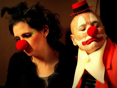 A fotografia mostra dois artistas caracterizados como palhaços, com maquiagem facial marcada e narizes vermelhos, posando com expressões sérias diante de um fundo neutro. A solenidade dos personagens contrasta com a expectativa de comicidade e reforça a ideia de que, por trás do riso, a palhaçaria também pode revelar vulnerabilidades e contradições humanas