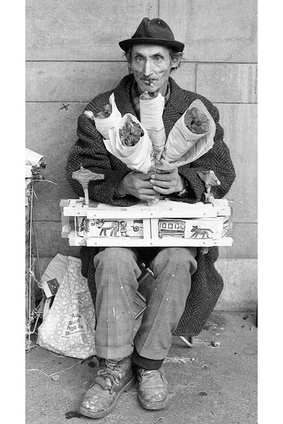 Photo by Eveline Meeuwse, Hans Krüsi (1920–1995) selling flowers at the Bahnhofstrasse in Zurich, with a “cow machine” on his lap, Zurich, Switzerland, 1978, Black and white photograph, 11.6 x 8.5" © Eveline Meeuwse