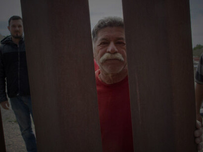 A Hispanic man in his sixties, with a mustache, is seen looking directly into the camera from the Mexican side of the steel-slat border wall between Mexico and the United States