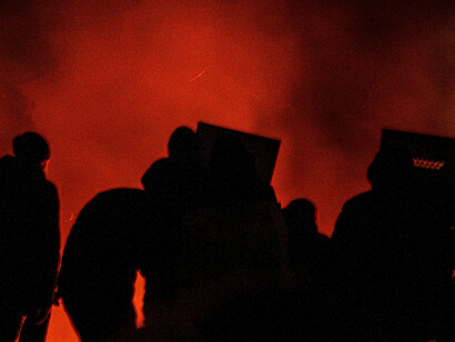 Silhouetted figures gather in a field at night, standing quietly behind police barricades under dim, distant lights