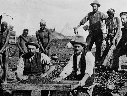 Black-and-white image of miners gathered around a diamond sorting table, with trays and tools, Kimberley, South Africa, 1800s