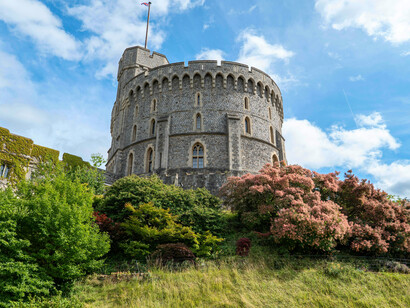 The Round Tower of Windsor Castle rises in gray stone beneath a clear blue sky, with the royal standard flying proudly at the summit, England