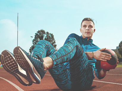 A male runner is engaging in leg stretches as part of his warm-up routine, preparing for a training run on the stadium tracks