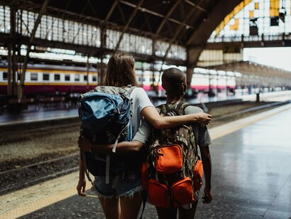 Vistas de costas, duas mulheres caminham juntas pela estação, cada uma levando sua mochila. Ter referências de companhia e segurança durante o trajeto pode tornar mais confortável a experiência de deixar para trás o ambiente familiar
