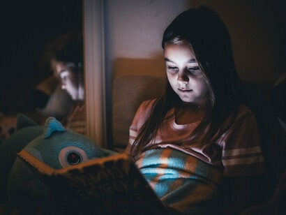 A young girl immersed in a book under the quiet glow of a light, representing the early spark of curiosity that begins a lifelong love of reading