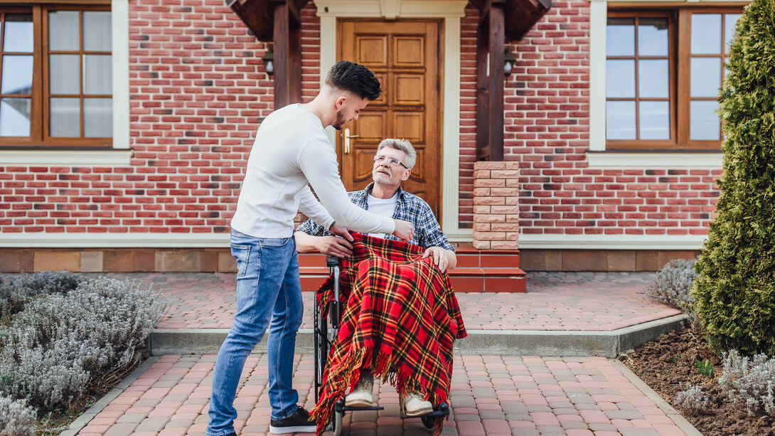 A man assisting an elderly person in a wheelchair in front of a house