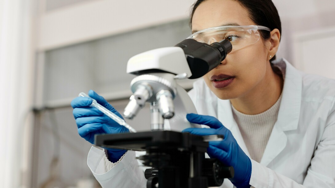 A woman wearing gloves and a lab coat looking at a sample through a microscope