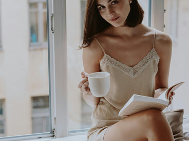 A woman sits by a window with a book and a cup of coffee, lost in quiet reflection as the morning light fills the room