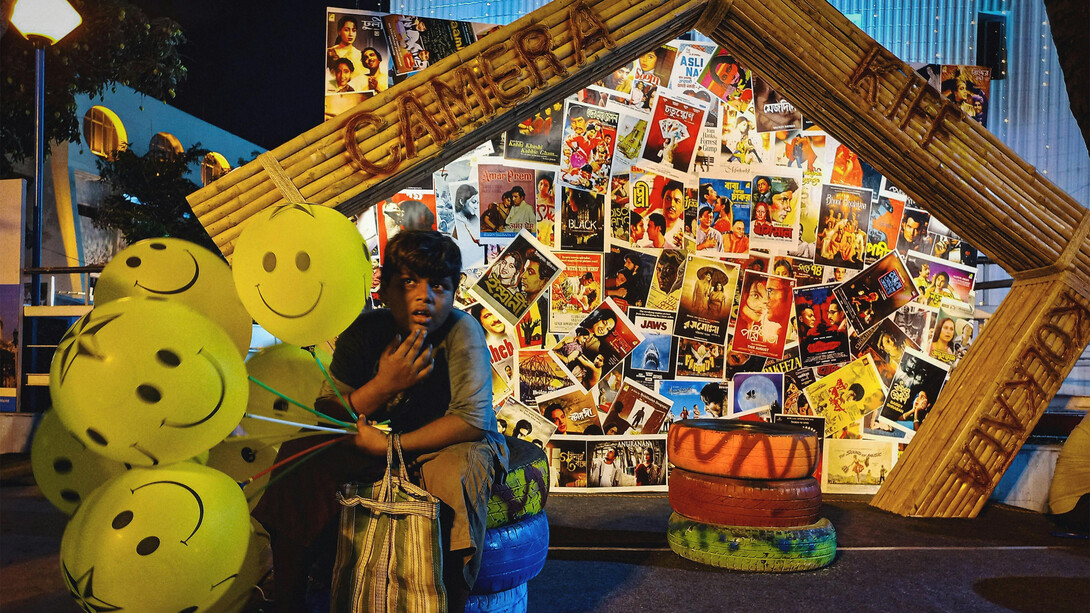 A child sits with balloons in front of a wall of movie posters, Kolkata, WB, India

