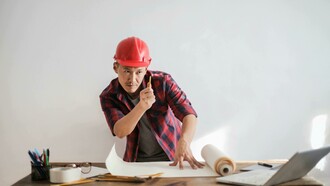A man in a red and black shirt wearing a red hard hat, an architect working with both computer and paper plans