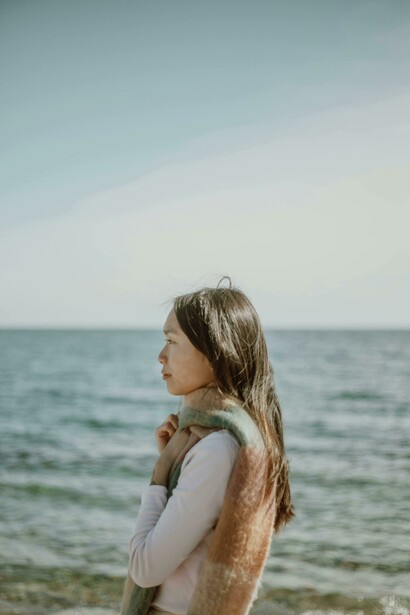 A serene Asian woman with long hair standing alone on the coast, quietly taking in the seascape and reflecting a sense of loneliness