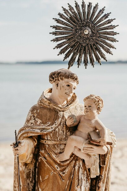 Em um enquadramento mais próximo, Santo Antônio aparece segurando o Menino Jesus, enquanto o mar compõe o cenário ao fundo. No romance A Cabeça do Santo, a devoção ao santo casamenteiro é transformada em elemento de uma história marcada por fé, desejos e encontros