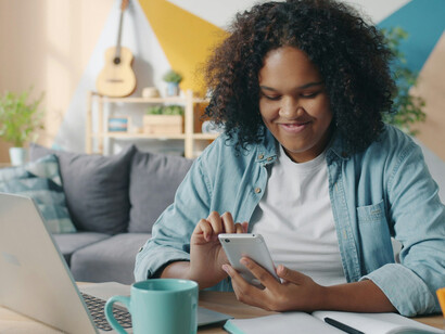 A young woman scrolling through her phone while working at a desk with her laptop