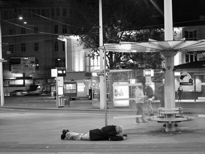 Um homem está deitado no chão em uma cena noturna registrada em preto e branco, nas proximidades de uma parada de ônibus. A imagem conduz à reflexão sobre as pessoas cuja vulnerabilidade permanece presente nos espaços públicos, mesmo quando passa despercebida