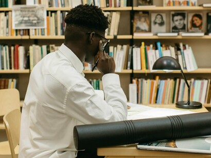 Jovem concentrado em sua pesquisa. O rapaz se aproximou do balcão central de atendimento e, após ter mostrado sua carteira da biblioteca, pediu os livros que havia reservado. Como alguns deles não poderiam ser emprestados, a simpática atendente levou-o até um dos grandes gabinetes de leitura, onde ele poderia ficar à vontade para consultá-los. Quando fosse sair, poderia levar consigo os demais exemplares que reservara



