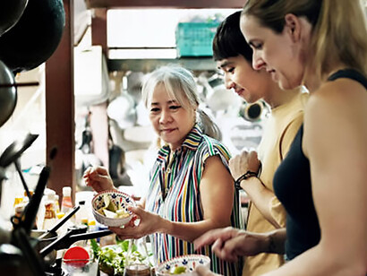 An experienced elderly woman demonstrates traditional Thai cooking techniques to tourists during a hands-on culinary class