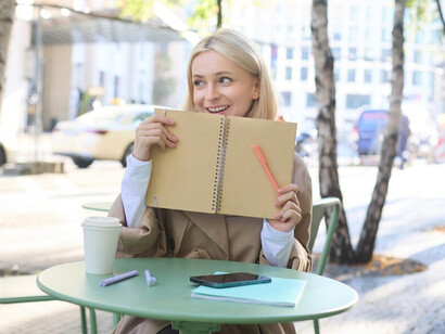 At a charming outdoor café, a young smiling blonde woman engages in journaling her gratitude while meditating in the fresh air