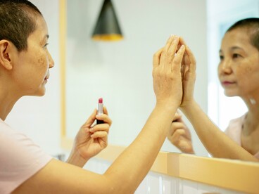 An Asian woman with breast cancer applies lipstick in front of a mirror, reclaiming a sense of normalcy
