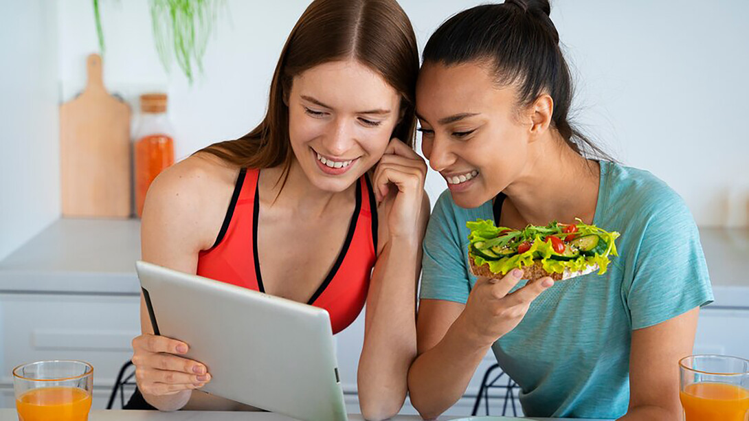 A woman in a kitchen, surrounded by healthy food, examines an online information source on the table