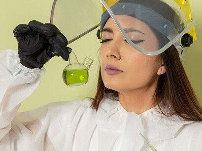 A female chemist in a protective suit holding a green solution on a green surface, observing its scent for testing purposes