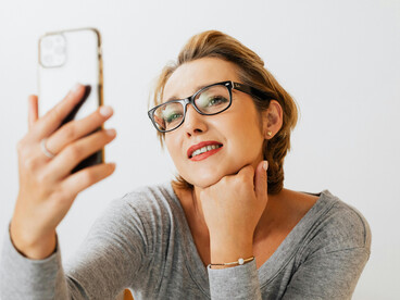 A woman in a gray long-sleeve shirt taking a selfie while repeatedly checking her phone