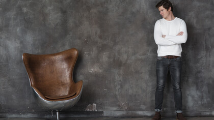 A man stands facing an empty leather armchair, appearing reflective and contemplative