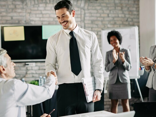 Um aperto de mão que simboliza reconhecimento e parceria, enquanto ao fundo a equipe celebra. Empresas que valorizam seus colaboradores constroem relações mais fortes, produtivas e alinhadas com o bem-estar coletivo