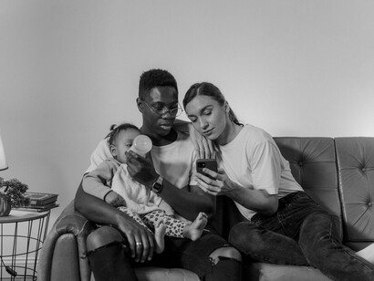O casal observa o celular enquanto alimenta a filha com uma mamadeira no sofá. O texto reflete sobre as mudanças nas relações familiares e os desafios da convivência na era digital