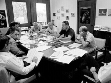 Centre spatial Johnson, 1982, à Houston, au Texas, États-Unis. Autour d’une table de débriefing, les astronautes des missions STS-2, STS-3 et STS-4 partagent leurs retours de vol. Au premier plan, John W. Young consigne les échanges, témoin attentif d’une nouvelle ère du programme spatial américain