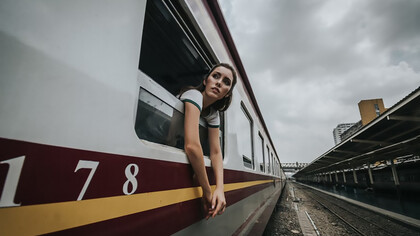 A woman’s head and hands are visible outside a train window under a cloudy daytime sky