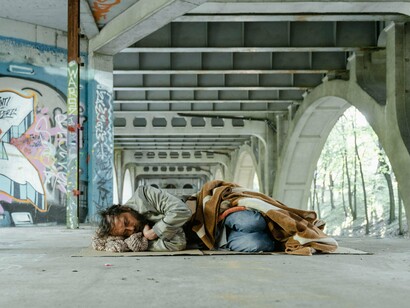 Um homem dorme sobre papelão sob um viaduto marcado por grafites. A cena revela a precariedade de quem utiliza o espaço público como abrigo e reforça a necessidade de uma sociedade capaz de reconhecer a dignidade humana para além das condições materiais
