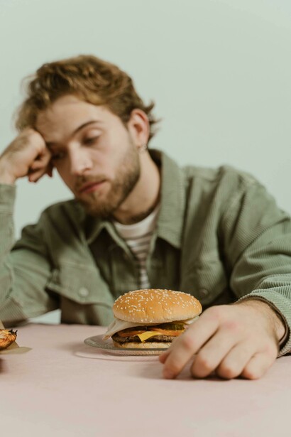 In a minimalist studio setting, a man sits with his head in his hand, gazing at a burger on the table, conveying a sense of depression