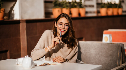 A young student with dark wavy hair, stylish glasses, and subtle makeup sits in a café journaling beside a cup of coffee, embracing a quiet moment of focus away from digital distractions