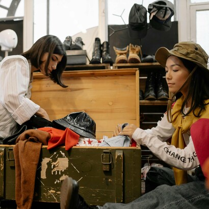 Friends examining the contents of a chest while trying on items they find, encapsulating the thrill of thrifting—an adventure in sustainability, self-expression, and the art of giving old clothes new stories