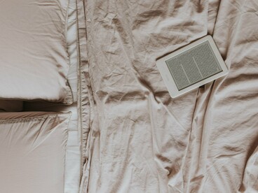 Um leitor de livros digitais ocupa o centro de uma cama coberta por tecidos claros, em uma composição marcada pela simplicidade e pelo conforto. A leitura contemporânea encontra no ambiente digital uma nova forma de preservar a experiência de mergulhar em uma história