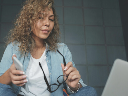 A woman wearing denim jeans works on her laptop while sitting on the bed
