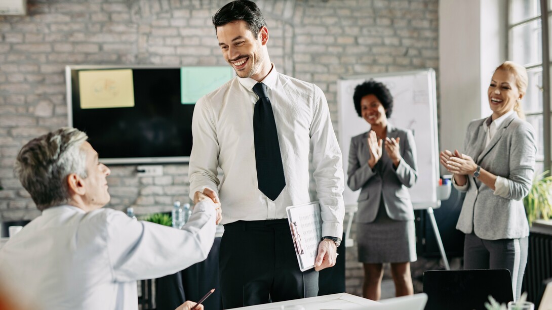 Um aperto de mão que simboliza reconhecimento e parceria, enquanto ao fundo a equipe celebra. Empresas que valorizam seus colaboradores constroem relações mais fortes, produtivas e alinhadas com o bem-estar coletivo