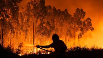 Silhueta de um homem segurando uma mangueira enquanto tenta conter uma floresta completamente tomada pelas chamas. A imagem simboliza a luta desigual contra um problema que se repete todos os anos sem soluções eficazes