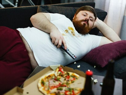 A man lying on the sofa, eating unhealthy snacks and mindlessly switching TV channels—a scene of a sedentary lifestyle