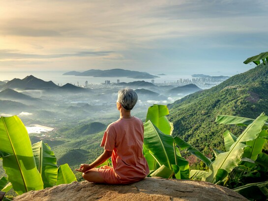 Uma mulher medita no alto da montanha enquanto o céu se dissolve entre luz, mar e cidade. Nem toda quietude visível significa silêncio por dentro. Sentada sobre a pedra, ela contempla o horizonte entre montanhas e o mar. Há respirações que atravessam paisagens inteiras sem conseguir alcançar a própria paz. Às vezes, o maior esforço da vida é simplesmente permanecer enquanto os pensamentos fazem barulho