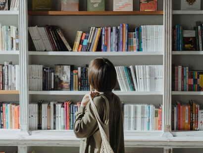 A woman in a beige long-sleeve shirt stands in front of a white wooden bookshelf