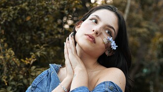 Uma mulher posa ao ar livre com flores delicadas presas ao rosto por um curativo transparente. Às vezes, os pequenos remendos que carregamos contam histórias maiores do que as marcas que procuram esconder. Tal como o botão extra, há gestos discretos que ajudam a manter intacto aquilo que a vida insiste em desafiar. Nem sempre a reparação elimina a marca, mas pode transformá-la em parte da história
