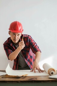A man in a red and black shirt wearing a red hard hat, an architect working with both computer and paper plans