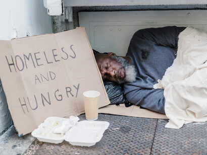 Un homme âgé dort sur un morceau de carton, la solitude et la vulnérabilité de la rue étant illustrées par une pancarte demandant de l’aide