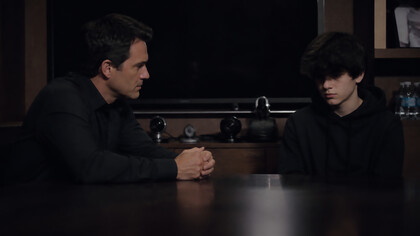 A father and son sit across from each other in silence, their tension shaped by control, performance, and the unseen weight of being watched