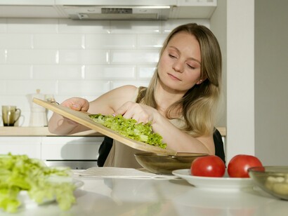 A woman in a beige tank top sitting in the kitchen near a table with vegetables