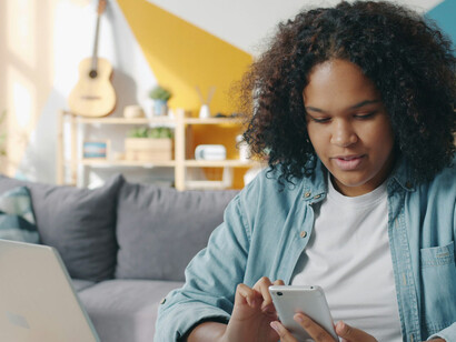 A young woman uses her phone at a desk, scrolling beside an open laptop