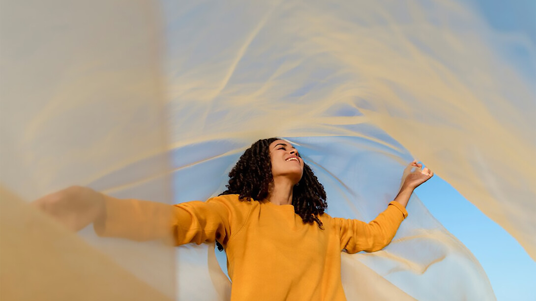 A woman stands in an open field, holding a flowing cloth that dances with the wind, symbolizing freedom and connection to nature