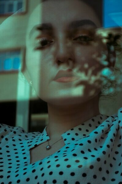 Behind a window, a woman in a blue and white polka dot shirt reflects quietly — a moment suspended between emotion and reality